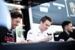 Three men sitting at a table signing autographs during an outdoor event with racing team branding.