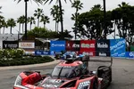 Red Acura race car number 60 racing on a palm tree-lined street circuit with banners in the background.