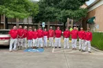 Group of 13 painters in red shirts and white pants posing outdoors near a tree and parked cars.