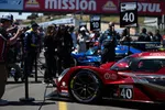 Red and blue race cars lined up on the grid with pit crew and officials preparing at a racing event under Mission and Motul banners.