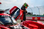 Race car driver in red and black suit with green helmet stands beside open door of Acura race car on track.
