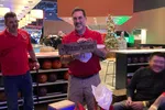 Happy man in red shirt holding a wooden gift plaque inside a festive bowling alley with Christmas decorations.