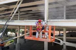 Worker in safety gear sandblasting or cleaning metal beams on building structure from elevated orange platform.
