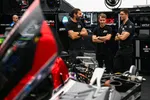 Three men in black shirts discuss near a partially disassembled race car in a professional racing garage.