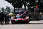 Red Acura race car with open door on racetrack surrounded by crew and blurred trees in background