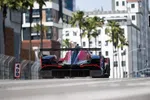 Rear view of a race car speeding on an urban street track with buildings and palm trees in the background