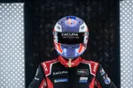 Race car driver in black Acura suit and colorful helmet standing between illuminated panels for dramatic effect.