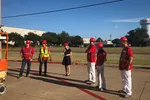 Group of six workers wearing red helmets and safety gear standing outside near construction equipment under clear blue sky.