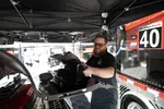 Bearded man wearing sunglasses working on car parts under a black canopy near a race truck marked 40