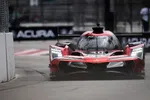Red Acura prototype race car with bright headlights racing on a street circuit near a concrete barrier.