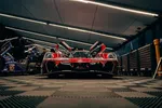 Front view of a red race car with open doors in a dimly lit garage surrounded by tools and crew members.