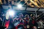 Racing team in matching red and black suits celebrating with fireworks in the background at night.
