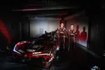 Two race car drivers in black and red suits stand beside a red Acura DEX race car in a dimly lit garage.