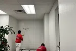 Two painters in red shirts applying white paint on office walls with rollers near a large green plant and ceiling lights.