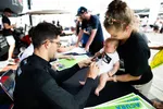 Man signing autograph for baby held by smiling woman at outdoor event with people in the background.