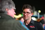 Man with glasses smiling and talking to another person at an outdoor event during nighttime.