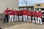 Group of construction workers wearing red safety vests, white pants, helmets, and face masks posing outdoors.
