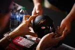 Person signing a black Bell racing helmet with autographs visible and bottles in the background.