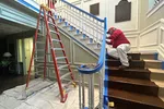 Worker in red shirt painting staircase railing in an indoor setting with ladder and protective sheets on floor.