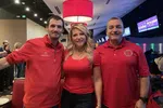 Three people smiling together in a bowling alley wearing red shirts under bright lighting.