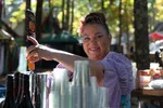 Smiling woman serving drinks outdoors with a flower in her hair and clear plastic cups stacked nearby.