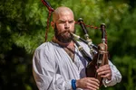 Bearded man in traditional attire playing bagpipes outdoors with lush green background on a sunny day