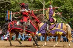 Two knights in red and purple armor jousting on horseback during a medieval reenactment event.