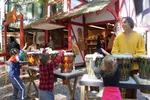 Children playing drums with an instructor at an outdoor festival booth with colorful buildings in the background
