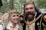 Smiling man and woman in ornate medieval royal costumes with crowns, pearls, and jewels at outdoor event.