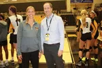 Two people smiling at a volleyball court with players in uniforms warming up in the background