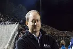 Man in black jacket standing near a sports field with a crowd in the background at night.