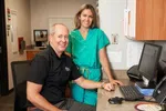 Smiling male doctor and female nurse in green scrubs at a medical office desk with computer and phone.