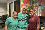 Three smiling medical staff celebrating a birthday with balloons and a banner in an office setting.