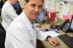 Male doctor in white coat working on computer in medical office with female colleague in background.