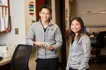 Two smiling people in gray fleece jackets standing in an office with books and a 3D spine model.