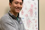 Smiling man in a gray jacket stands in front of an anatomical hand and wrist chart on a beige wall