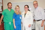 Group of four healthcare professionals smiling indoors wearing scrubs and a white coat in medical office.