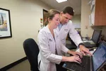Two medical professionals in white coats working together on a laptop in a clinical office environment.