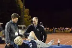 Two men attend to a young male athlete lying on a table at night on a running track during an outdoor sports event.