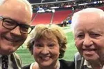 Three smiling older adults taking a selfie at an indoor stadium with a green field and red seats in the background