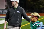 Football coach in black jacket and white cap supports seated player with blonde hair and maroon headband on field