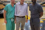 Three men standing on a gymnasium floor, two in medical scrubs and one in casual business attire, smiling at camera