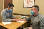 Doctor wearing a mask examining patient's wrist in a medical office with anatomy chart on the wall