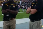 Two football coaches stand with arms crossed on the sidelines of a green football field during a game.