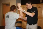 Man in black shirt demonstrating arm technique to another man in gray Adidas shirt in a sports locker room