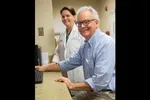 Male patient and female doctor at a medical office using a computer, smiling towards the camera.