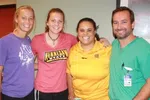Group of four smiling people standing together, including two women in casual shirts, one in yellow polo, and a man in green scrubs.