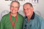 Two older men smiling, one in a green shirt with a red lanyard, the other in a blue shirt, against a branded backdrop