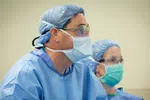 Two medical professionals wearing surgical masks, caps, and gowns in a clinical setting focused on patient care.