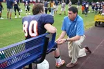 Football coach kneels talking to player in uniform sitting on bench during practice on grassy field.
